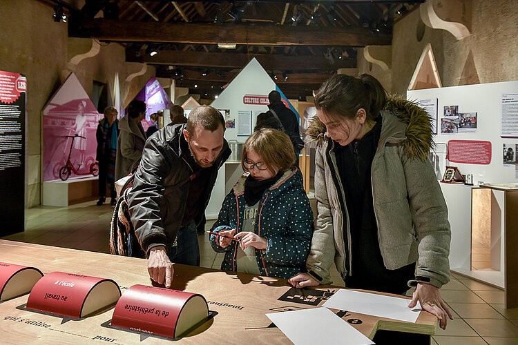 Un père et ses deux filles regardent un objet sur une table que le père montre du doigt. Dans le fond, une exposition - Agrandir l'image, fenêtre modale