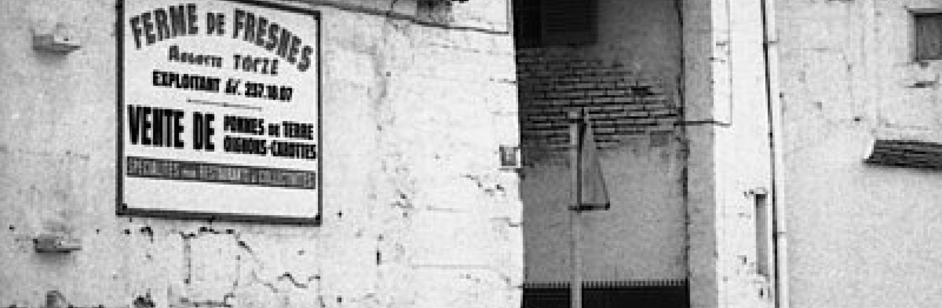 Photo noir et blanc probablement prise dans les années 70, on distingue l'entrée de la ferme derrière des voitures et un panneau indique : "Ferme de Fresnes" "Vente de pommes de terre, oignons, carottes"