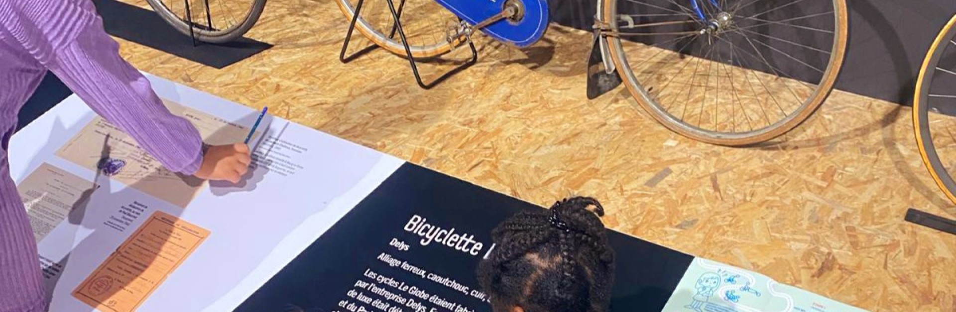Trois enfants devant un vélo de collection remplissant un livret de chasse au trésor, dans la grande salle d'exposition de l'écomusée