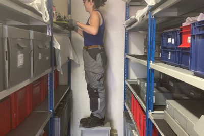 Chargée des collections rangeant des objets entourée des rayons des réserves - Agrandir l'image 1 sur 3, fenêtre modale