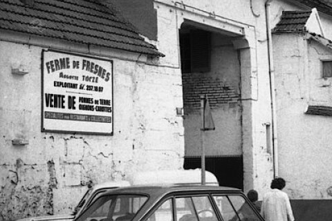 Photo noir et blanc probablement prise dans les années 70, on distingue l'entrée de la ferme derrière des voitures et un panneau indique : "Ferme de Fresnes" "Vente de pommes de terre, oignons, carottes"