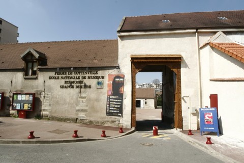 Entrée d'une ancienne ferme qui laisse apercevoir une cour et des immeubles en fond.