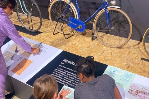 Trois enfants devant un vélo de collection remplissant un livret de chasse au trésor, dans la grande salle d'exposition de l'écomusée